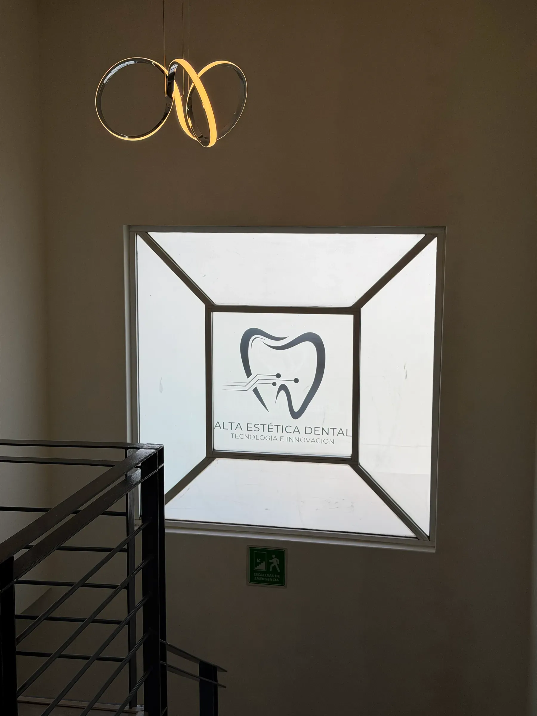 Ventana con el logotipo de Alta Estética Dental iluminado sobre la escalera, bajo una lámpara colgante.