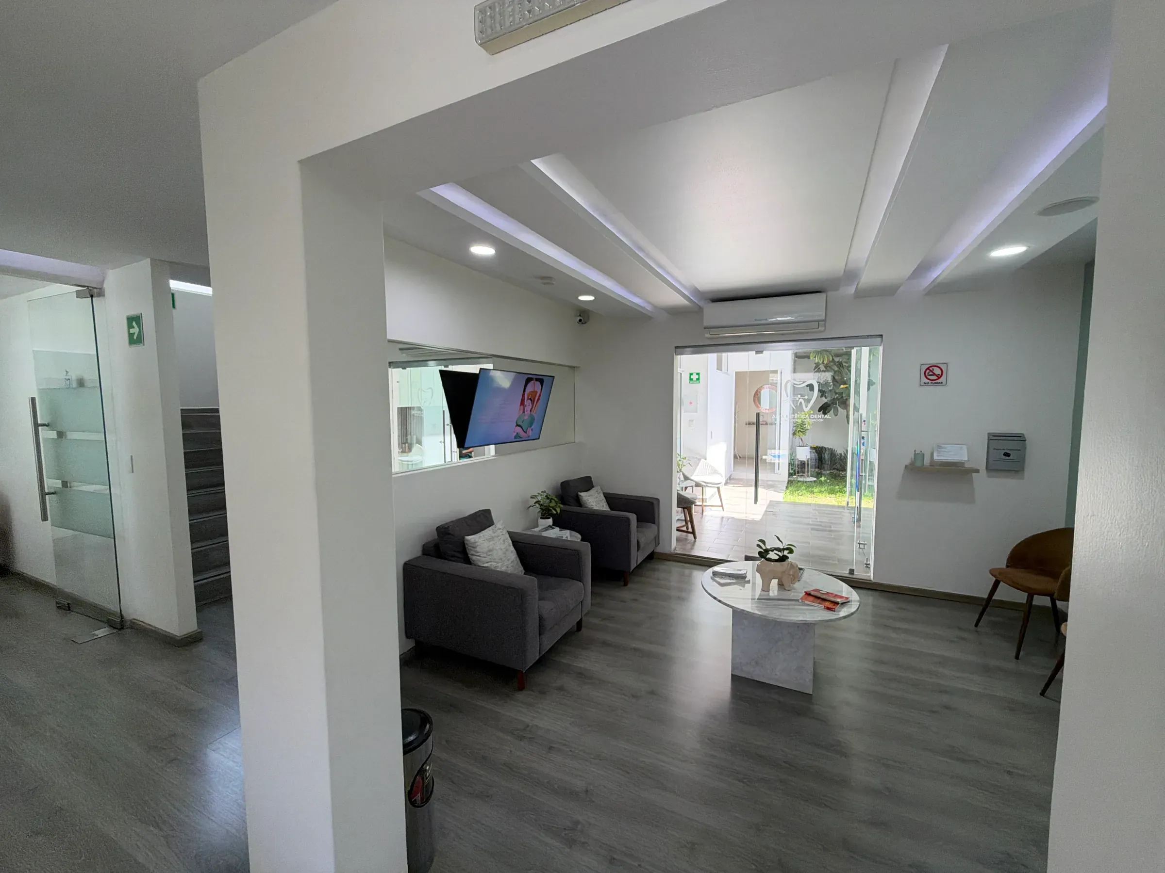 Sala de espera con sillones grises, mesa de centro y pantalla, con luz natural al fondo.