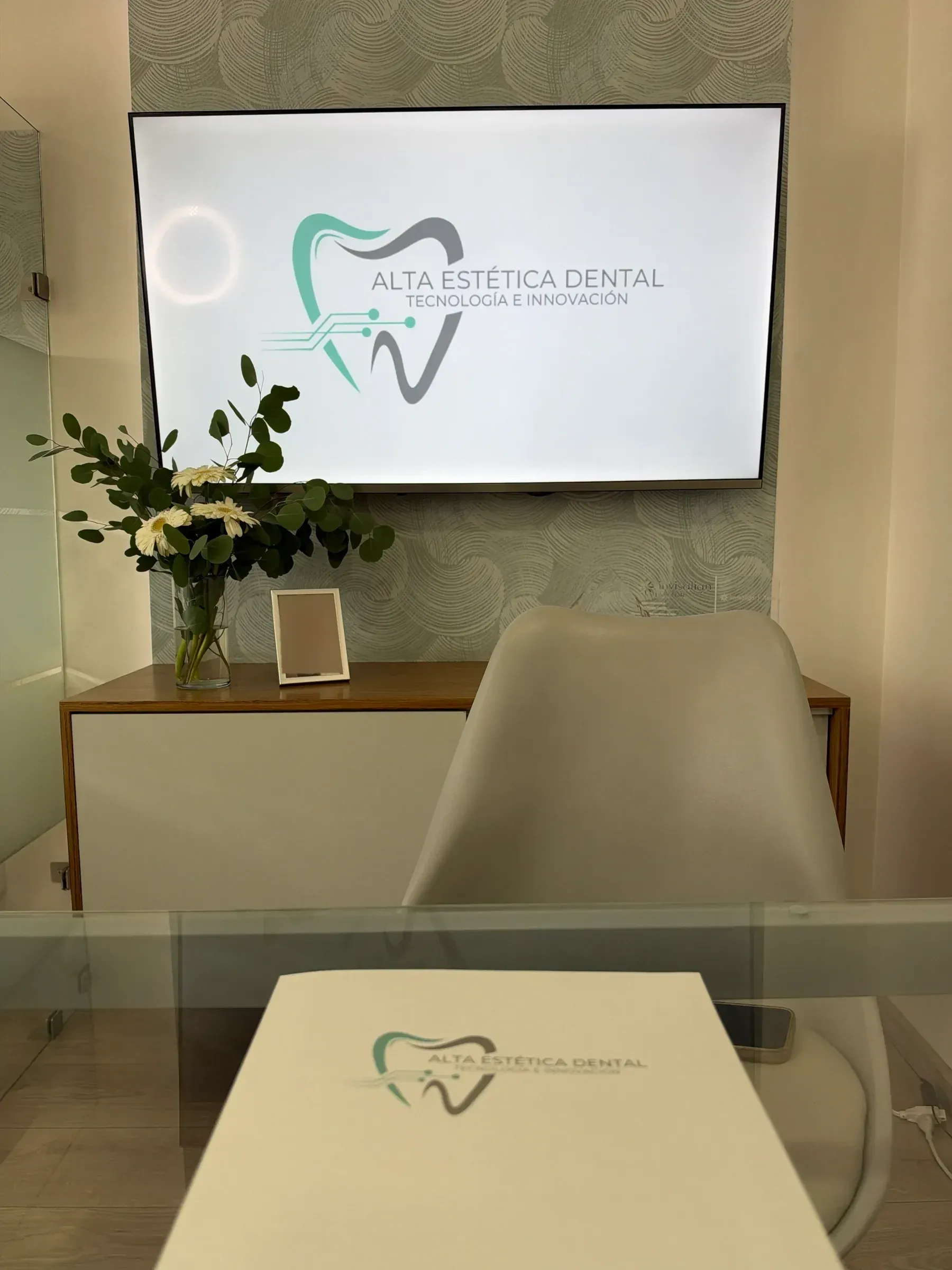 Escritorio de cristal frente a una pantalla con el logotipo de la clínica, con flores blancas.